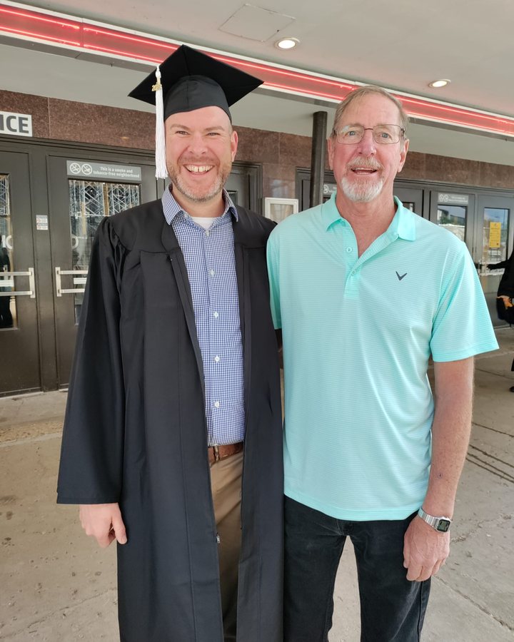 Aaron in a graduation cap and gown, smiling beside his dad, Dave