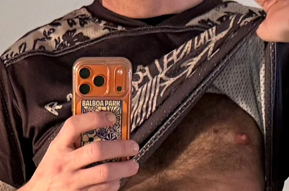 Aaron Berth in a mirror selfie, face out of frame, shirt lifted, phone in an orange Balboa Park case