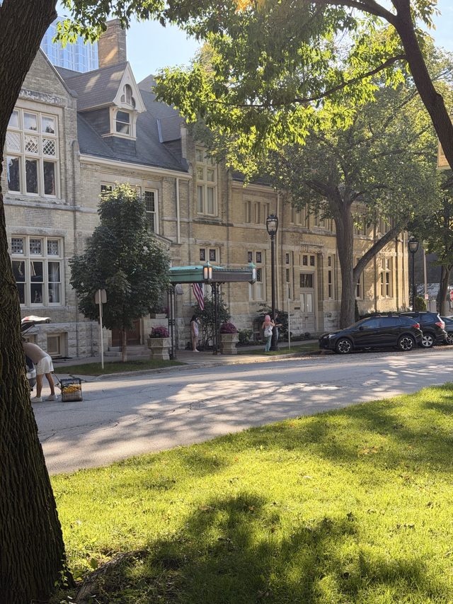 The Athenaeum, a stone Victorian clubhouse with a green iron entrance canopy, seen across a sunny lawn through the trees.