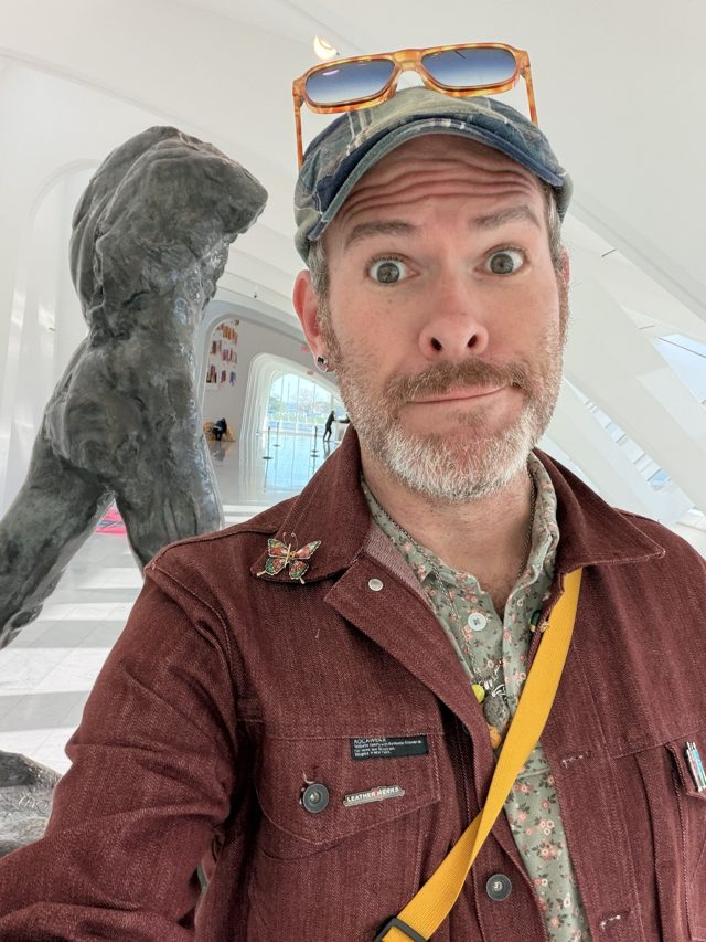 Aaron, eyebrows up, in a cap and red-brown chore jacket inside the white ribs of the Calatrava hall.