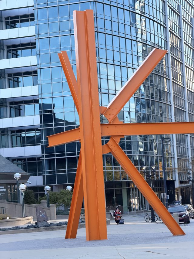The Calling, orange steel I-beams bolted into a star, in front of a glass office tower.