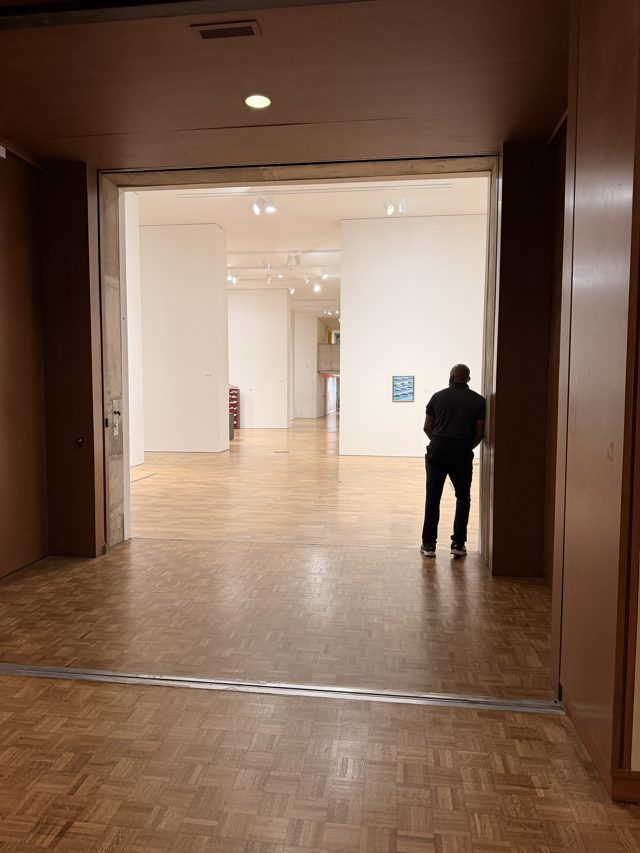 From a dark wood doorway, a bright white gallery; a man in dark clothes leans in the far doorway, seen from behind.