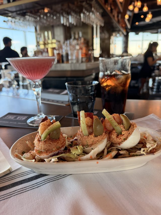 Bloody Mary deviled eggs topped with shrimp, celery, olives and bacon on slaw, the pink drink behind.
