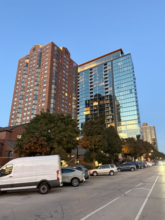 A brick tower and a glass tower at dusk, lit windows, parked cars along the street.