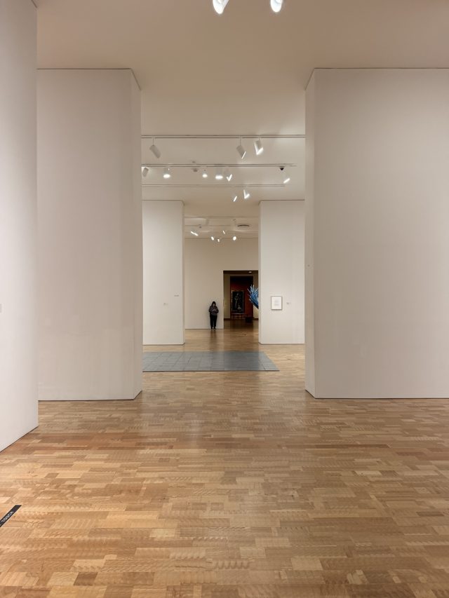 White gallery walls stepping back in a long corridor to a red room with a painting at the end.