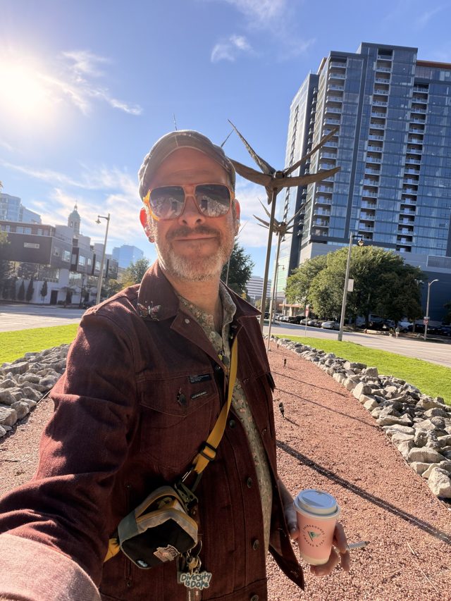 Aaron in sunglasses with a coffee, a tall bronze bird sculpture with outstretched wings behind him, towers beyond.