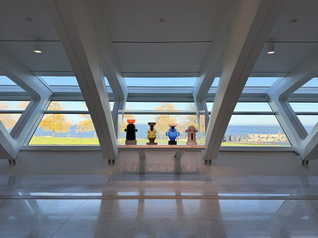 Four colored glass vessels in a case before slanted white beams and windows onto yellow trees and the blue lake.