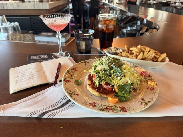 A crisp cutlet under a heap of greens on a flowered plate, fries behind, the notebook open beside it mid-poem.