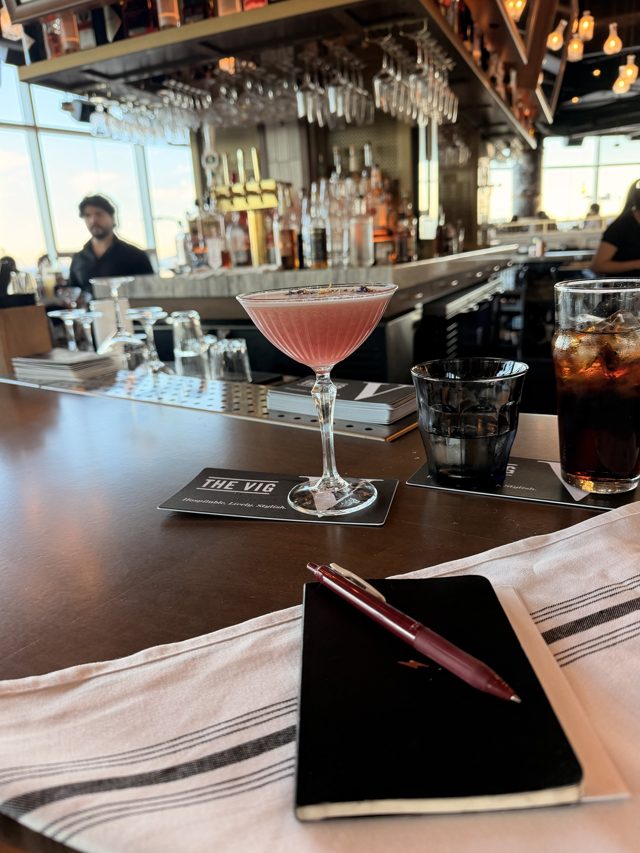 A pink Sour Grape in a coupe on a The Vig coaster, a black notebook and red pen on a striped towel in front.