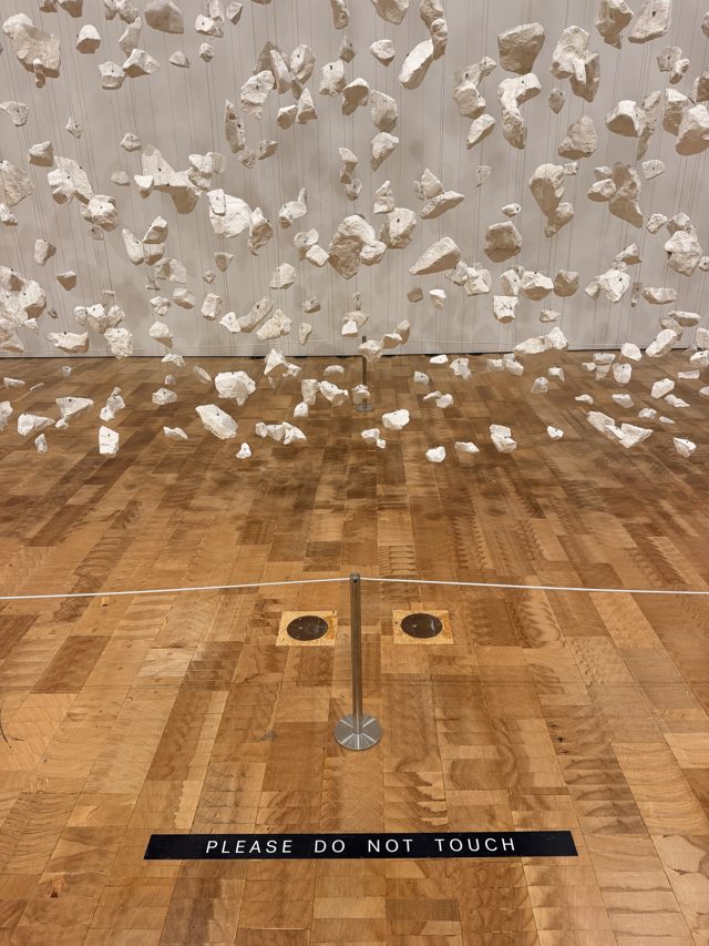 Hundreds of white stones hung in the air over a parquet floor, a small sign reading Please do not touch.