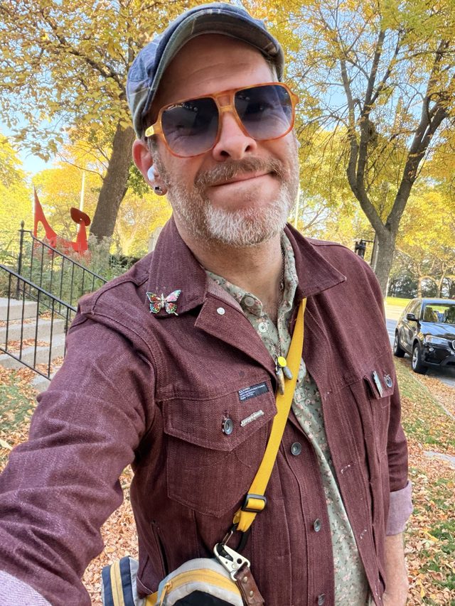 Aaron grinning in orange sunglasses under yellow autumn trees, the orange beams of The Calling behind his shoulder.