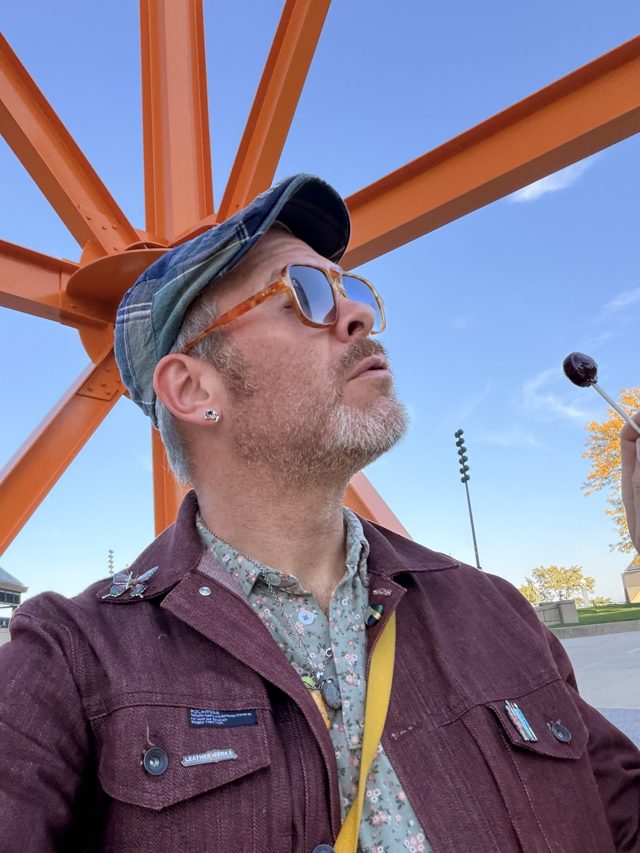 Aaron in a plaid cap and orange sunglasses eyeing a tootsie pop
