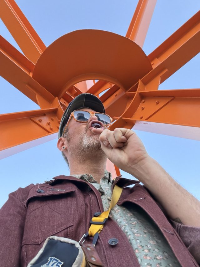 Looking up at Aaron biting a tootsie pop, The Calling’s orange hub and beams against blue sky above him.