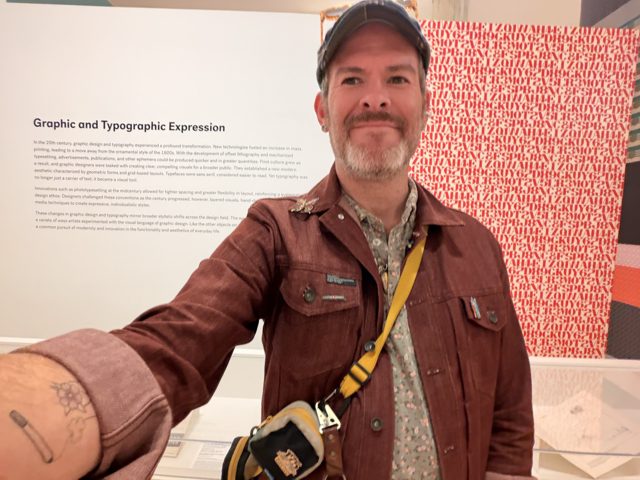 Aaron smiling in a cap beside a gallery wall titled Graphic and Typographic Expression and a red wall of stencil letters.