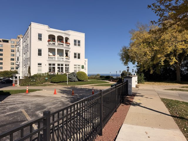 White Manor, a white apartment house with balconies, set back behind an iron fence with the lake beyond it.
