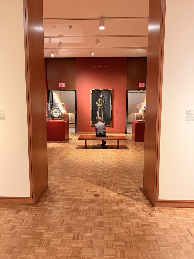 Through a doorway, a red gallery wall with Zurbarán’s Saint Francis and one visitor on a bench, seen from behind.