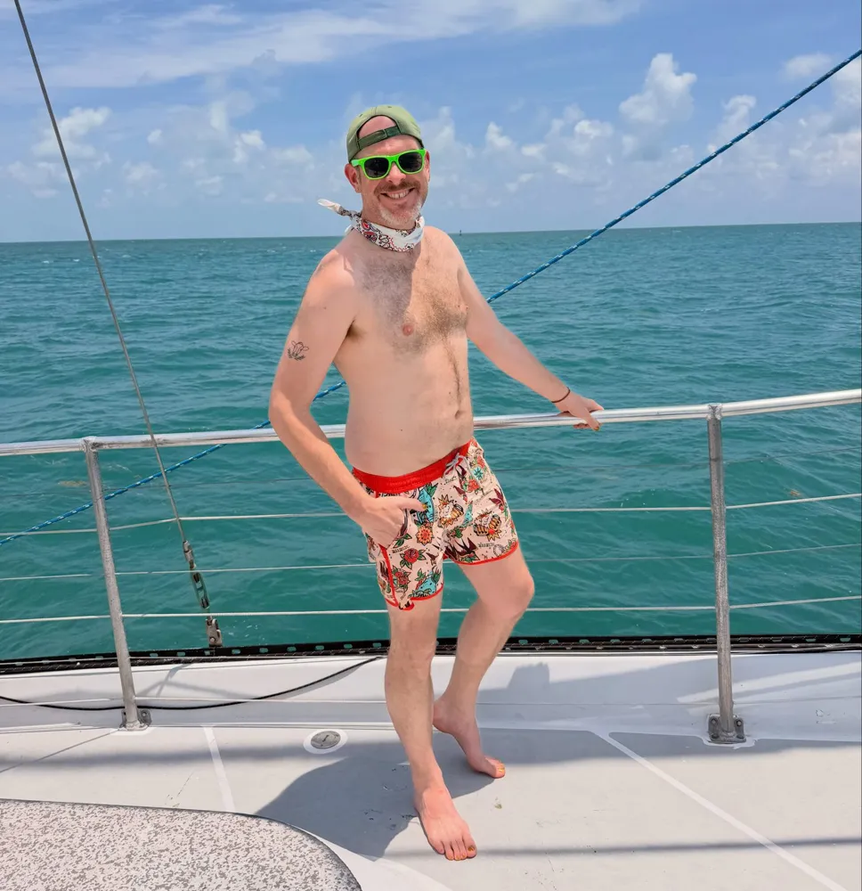 Aaron in red swim trunks posing at the bow of the sailboat, open water behind