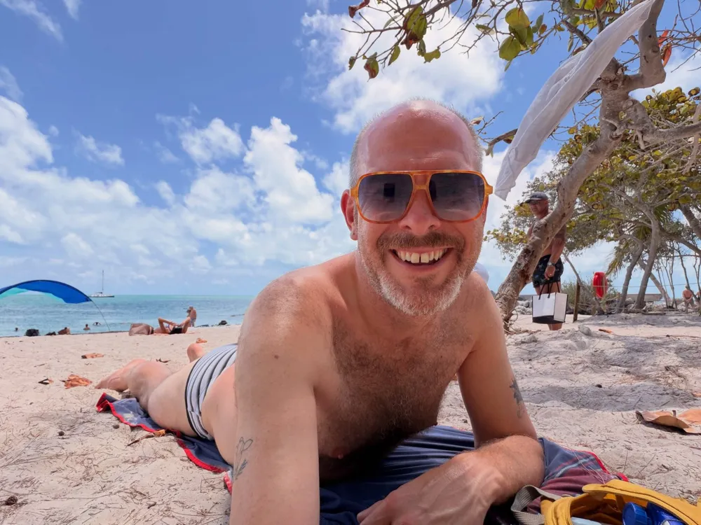 Aaron in a swim brief lying on a towel on the beach, grinning