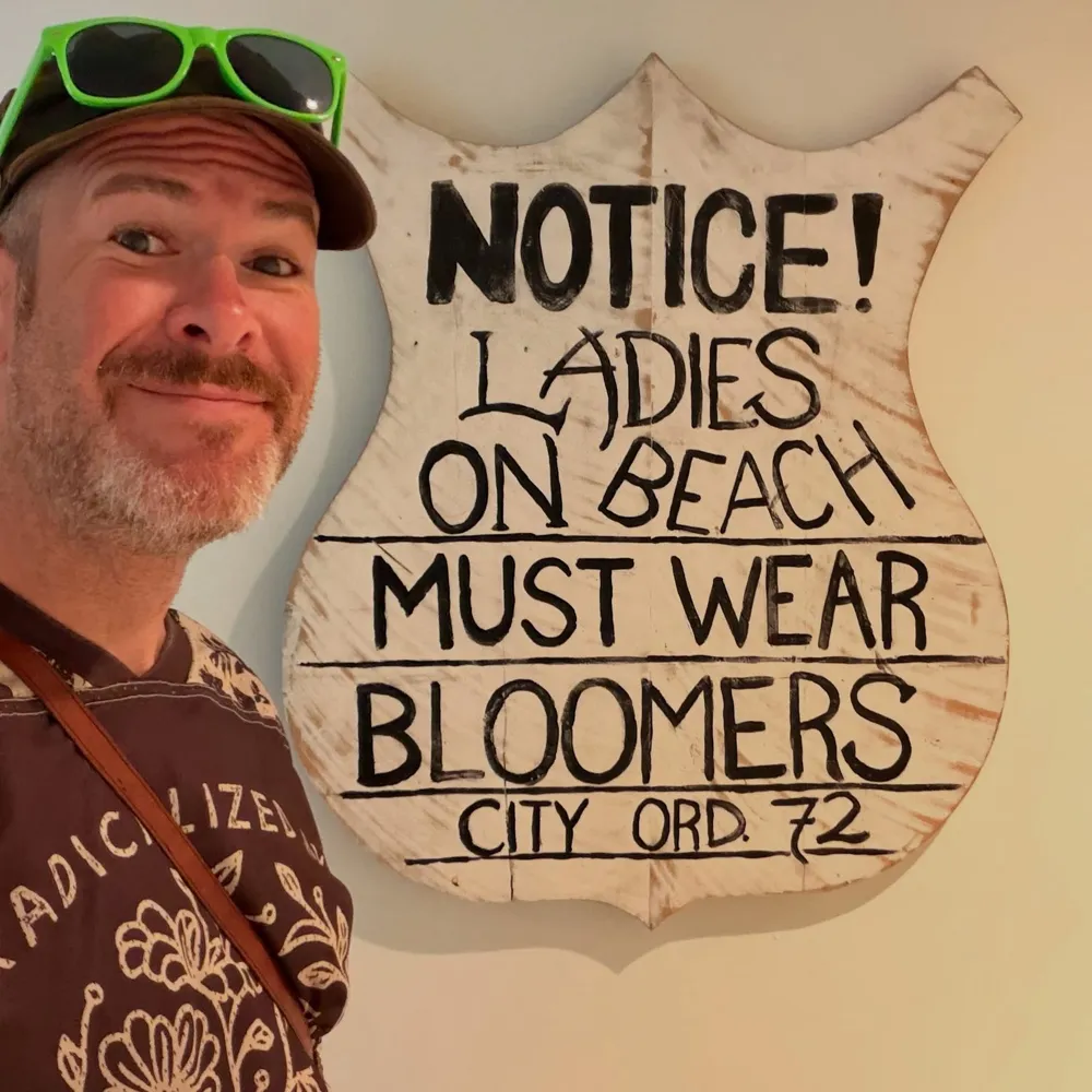 Aaron beside a sign reading Notice! Ladies on beach must wear bloomers, City Ord. 72