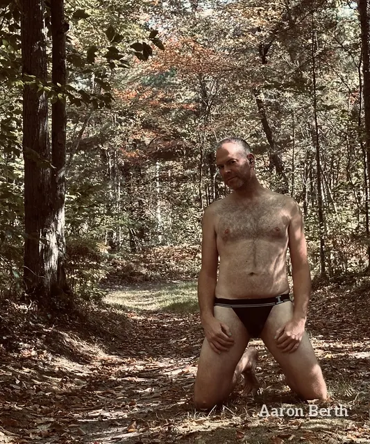 Aaron kneeling on a leafy forest path in black briefs, looking at the camera