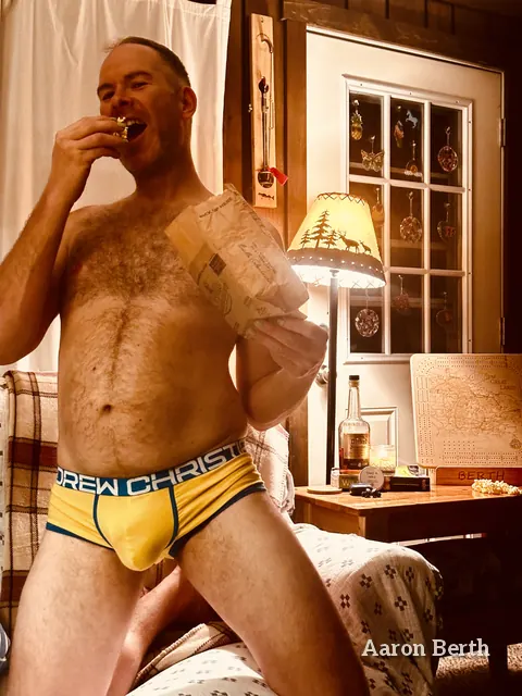 Aaron in a cabin in yellow briefs, eating a snack by a lamp and a window