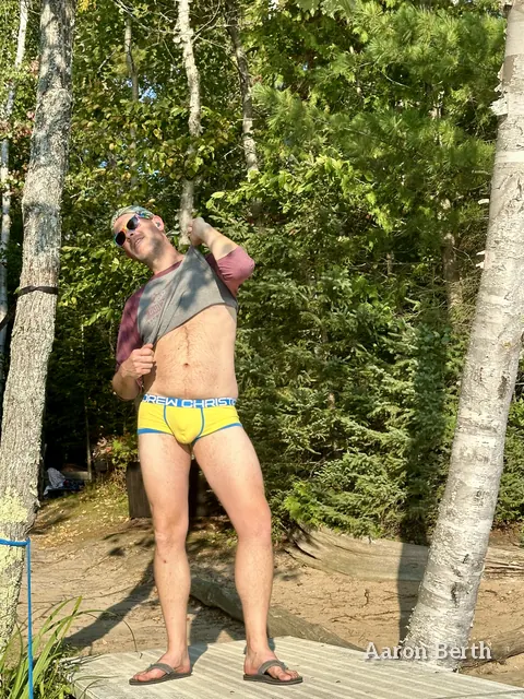 Aaron on a lake dock in yellow briefs, lifting a plaid shirt, with sunglasses and a cap