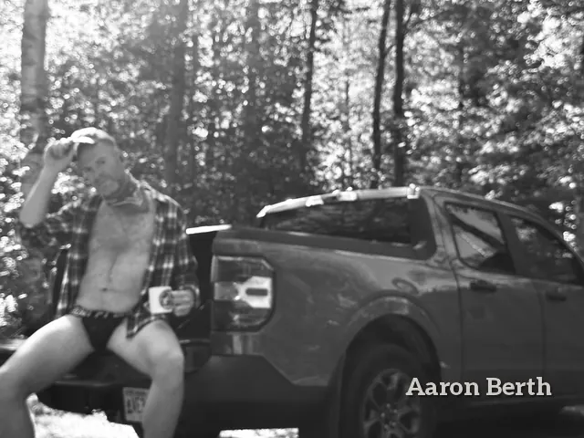Black and white: Aaron leaning on a pickup truck in an open plaid shirt, holding a mug