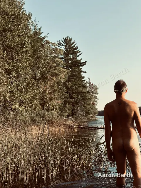 Aaron from behind at a lake edge, nude, looking out over the water and pines