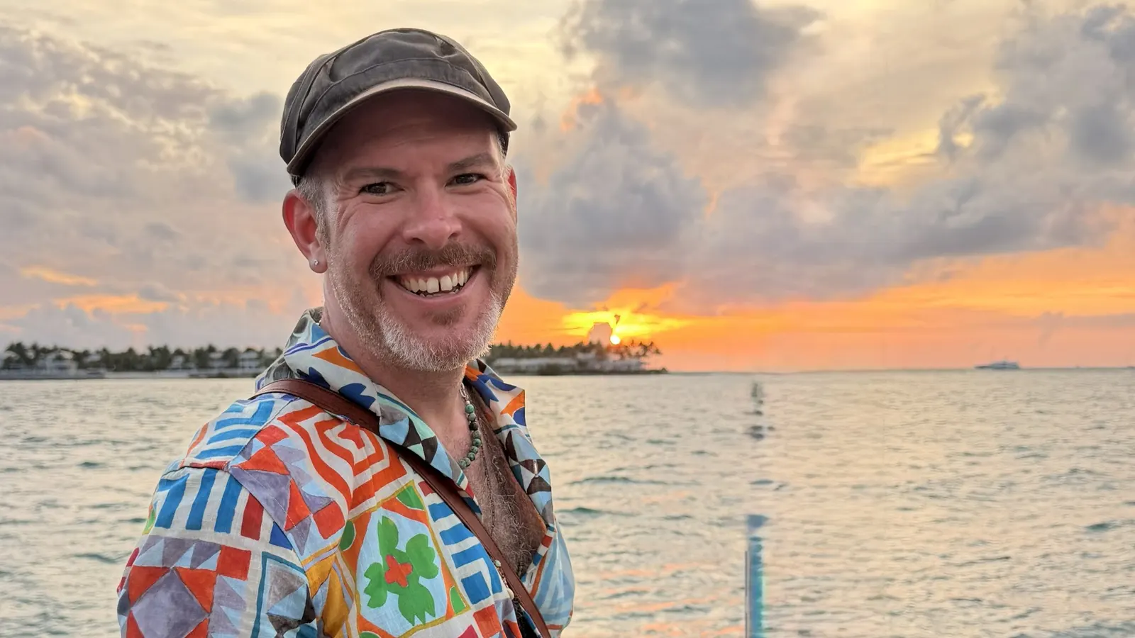 Aaron Berth smiling in a bright patterned shirt at a sunset on the water