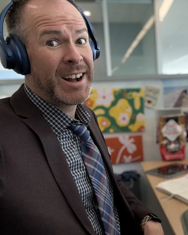 Aaron in headphones and a blazer, mid-call at his desk, eyebrows up